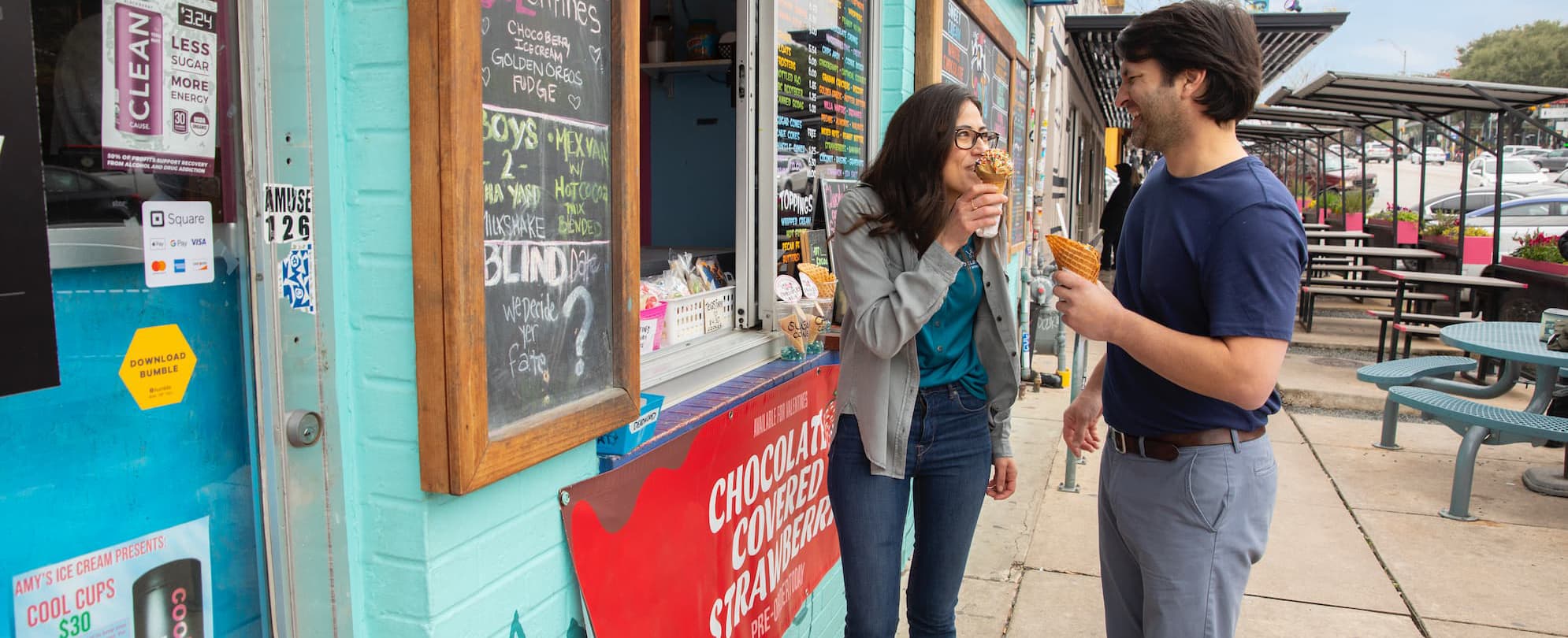 A smiling man and woman enjoy ice cream cones while standing in front of the store window.