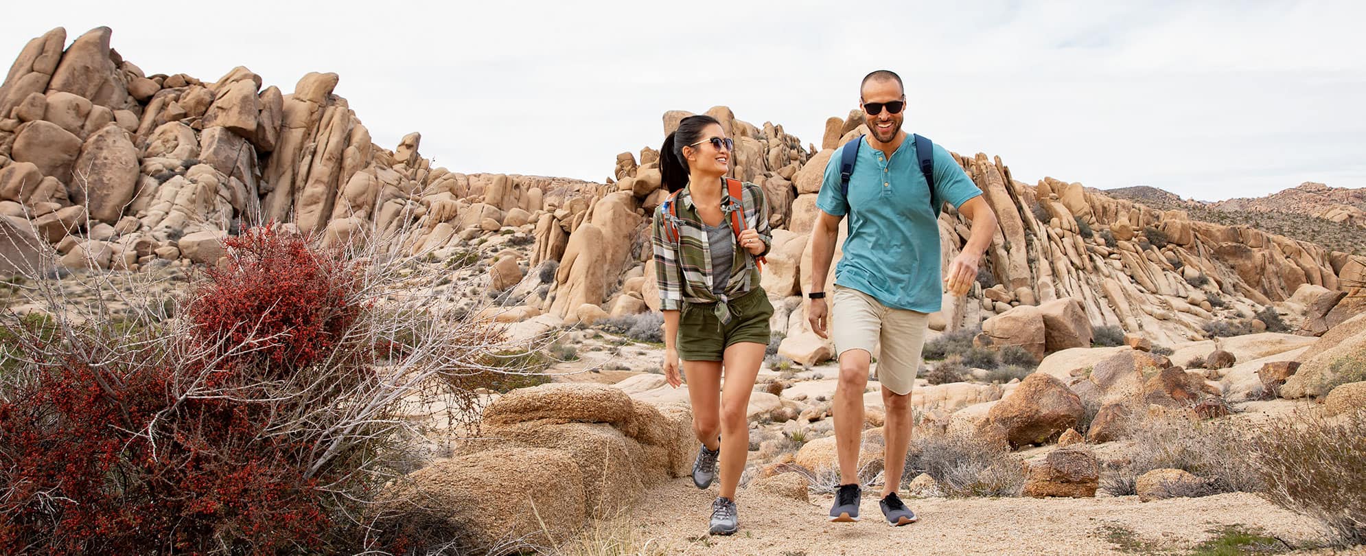 Smiling man and woman wearing sunglasses and backpacks hike along rocky path during their WorldMark by Wyndham vacation.